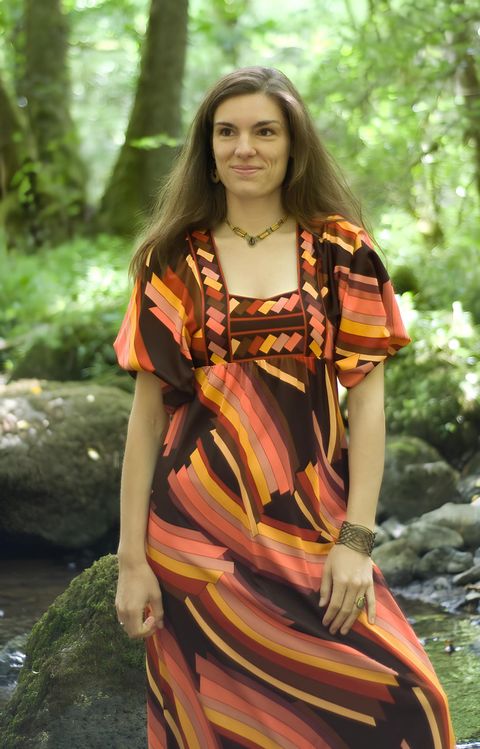 Une femme se tient dans une forêt verte à côté d'un ruisseau. Elle porte une robe colorée et à motifs.