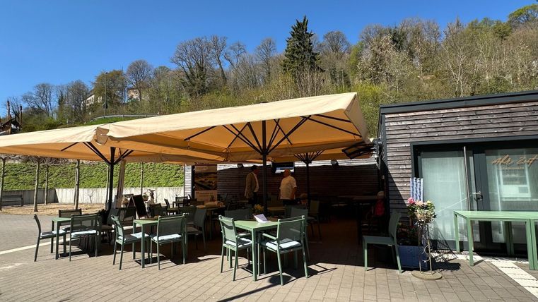 Ein gemütlicher Außenbereich eines Restaurants mit grünen Tischen und Stühlen. Im Hintergrund sind Sonnenschirme und eine grüne Landschaft zu sehen.