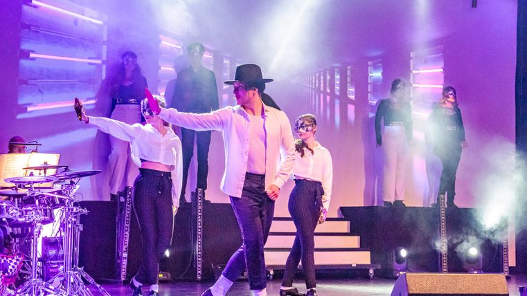 Eine Gruppe von Tänzerinnen in eleganten Kostümen präsentiert eine Show auf der Bühne. Im Hintergrund sind Lichter und ein großes Zeichen zu sehen.