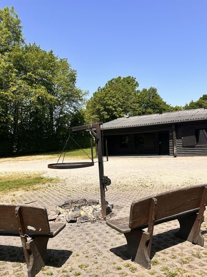 Een rustige plek in de buitenlucht met twee banken en een kampvuurgebied. Op de achtergrond staat een houten gebouw en de zon schijnt.