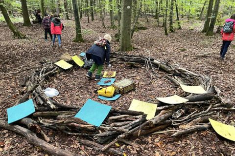Eine Gruppe von Menschen arbeitet im Wald an einem kreativen Projekt. In der Mitte sieht man einen Kreis aus Ästen mit bunten Papierblättern.