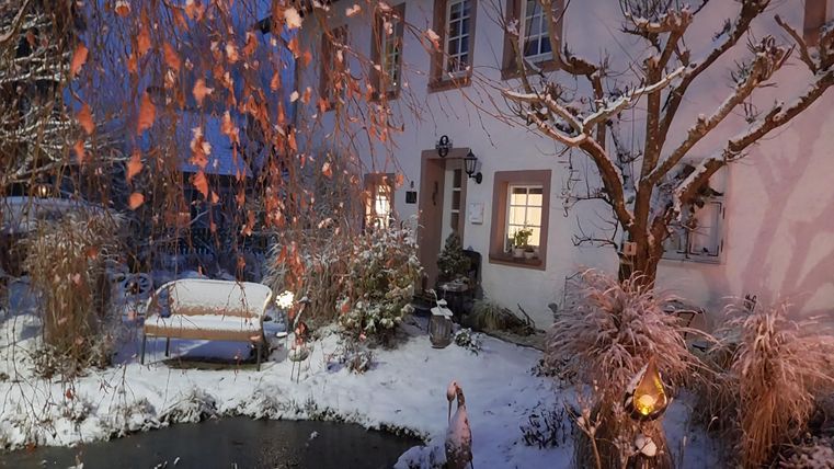 A pretty, snow-covered house at dusk. The garden is atmospheric with a small pond, a bench, and warm lights decorated.