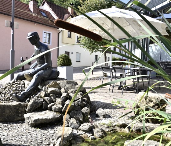 Statue en bronze d'un homme sur des pierres, entouré de roseaux et d'un petit étang. En arrière-plan, on aperçoit des maisons et un parasol., © Tourist-Information Bitburger Land