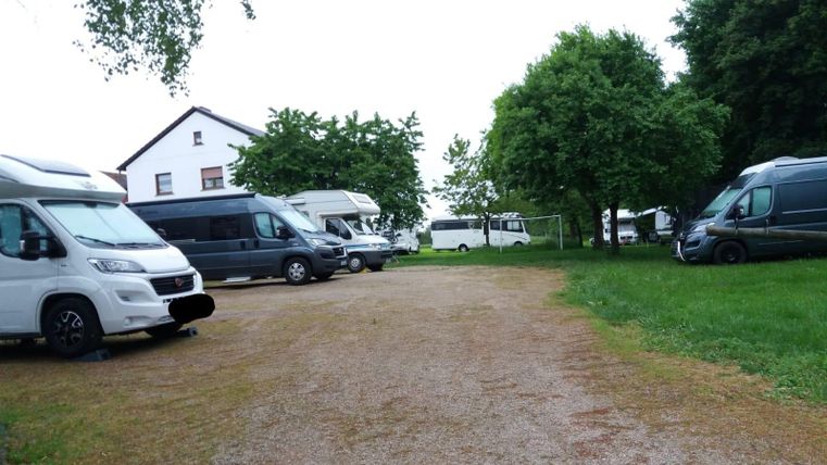 Motorhome parking area with several parked motorhomes, surrounded by trees and a white building in the background.