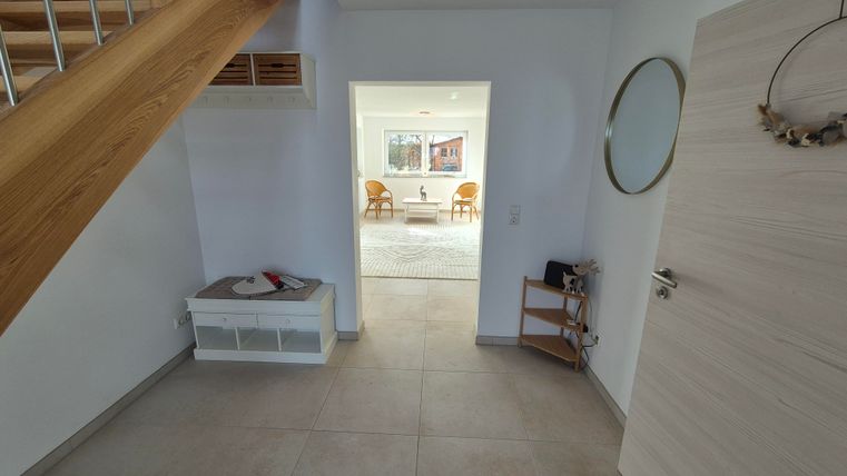 Une entrée lumineuse avec un escalier en bois. À travers la porte ouverte, une autre pièce avec des meubles est visible.