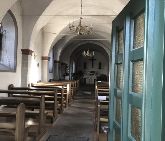 Vue d'une petite chapelle avec des bancs en bois, un autel et des chandeliers. La porte verte est ouverte, la lumière passe à travers les fenêtres., © TI Bitburger Land - Steffi Wagner