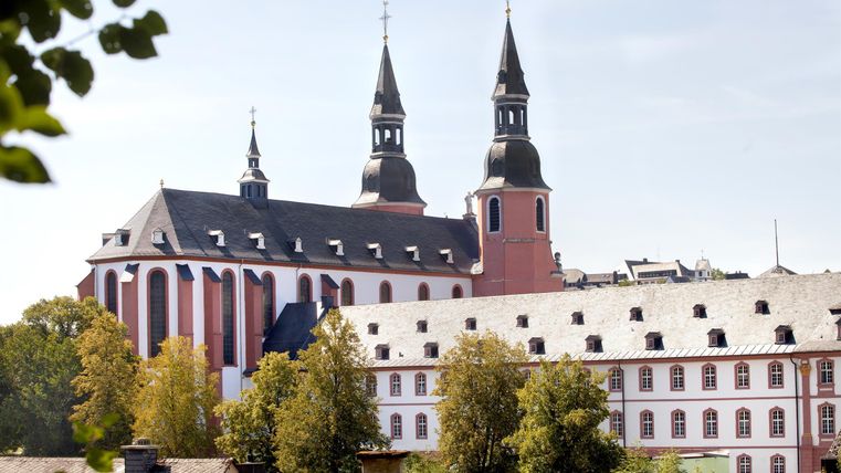 An impressive church with two tall towers and a charming facade. Surrounded by green trees and a clear sky.