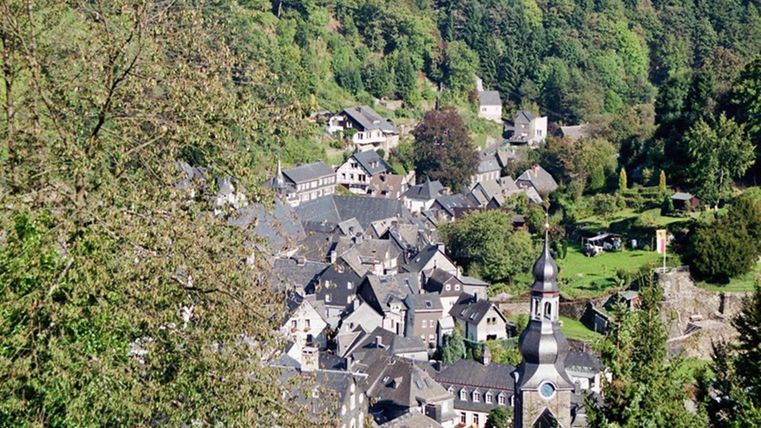 Un village pittoresque dans une vallée, entouré de forêts verdoyantes. Les toits des maisons sont gris foncé, et à l'arrière-plan, une église est visible.