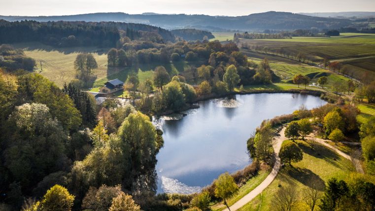 Vue aérienne d'un lac dans la petite vallée de Bolsdorf près de Hillesheim, entouré d'arbres et de champs.
