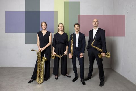 Eine Gruppe von vier Musikern steht vor einer minimalistischen Wand. Sie halten Saxophone und tragen elegante, schwarze Kleidung.