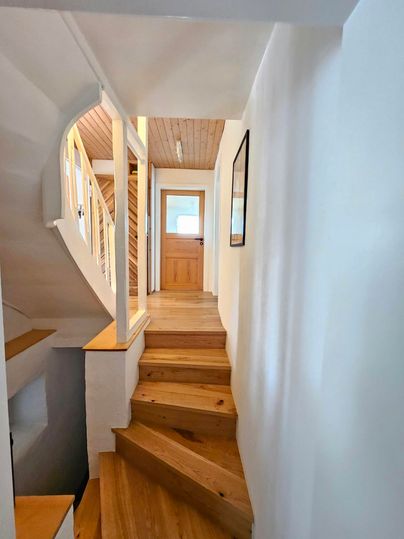 Een lichte gang met een houten trap die naar een deur leidt. De muren zijn in neutrale kleuren gehouden, en het licht stroomt van de opening boven.