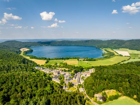 Panorama-Luftaufnahme des Laacher Sees, umgeben von dichten Wäldern und Feldern; im Vordergrund liegt die Abtei Maria Laach mit ihren Gebäuden eingebettet in die grüne Landschaft unter blauem Himmel mit vereinzelten Wolken.