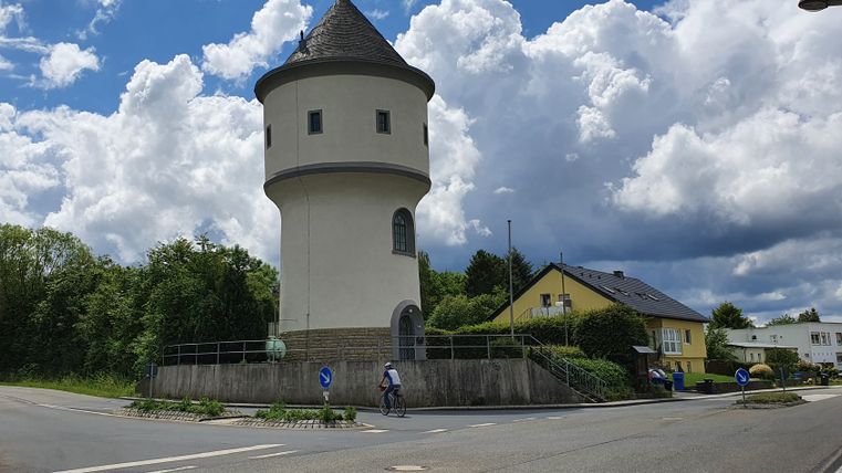 Un château d'eau rond au toit pointu se trouve à côté d'une route, entouré d'arbres et de maisons. Un cycliste passe, le ciel est nuageux.
