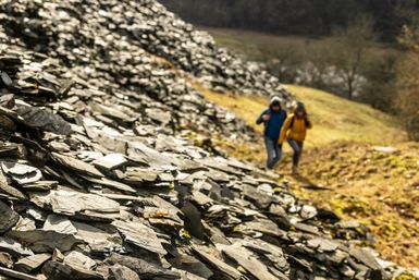 Zwei Wanderer gehen auf einem schmalen Pfad am Rand eines großen Schieferhaldenhangs im Kaulenbachtal entlang; die Landschaft ist hügelig und von Natur geprägt, im Hintergrund Wiesen und Wälder bei weichem Licht.