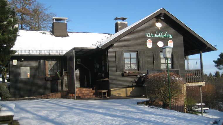 Ein schwarzes Holzhaus mit weißen Fenstern liegt im Schnee. Der Himmel ist klar und blau.