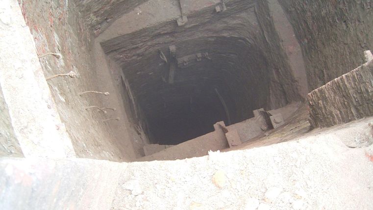 A dark shaft with steep walls and few visible steps. The floor is covered with loose material.
