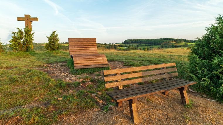 Een houten bank en een ligstoel staan op een heuvel met uitzicht op het landschap. Op de achtergrond zijn bomen en een kruis te zien.