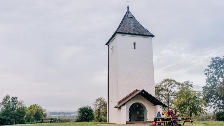 A white tower with a pointed roof stands on a meadow, with people sitting at a picnic table next to it.