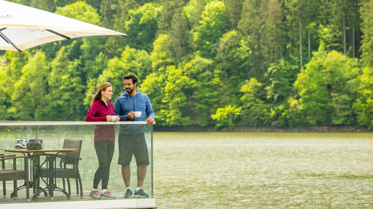 A woman and a man with a cup in their hands stand on the terrace of a café by the water