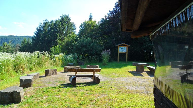 Un endroit paisible dans la nature avec une prairie et des bancs en bois. À l'arrière-plan, des arbres et un panneau d'information sont visibles.