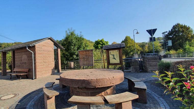Place du village de Brecht avec banc circulaire et panneaux d'information, entourée d'arbres et d'un petit bâtiment.