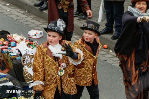 Two children in distinctive costumes with top hats are standing on a street. They are happily posing and enjoying a festive atmosphere.