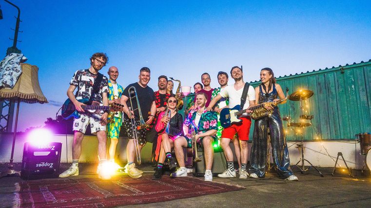 Un groupe coloré de musiciens se tient sur une scène en plein air. Ils portent des vêtements voyants et jouent divers instruments au coucher du soleil.
