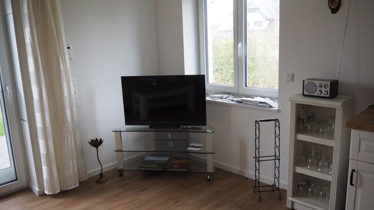 Ein gemütlicher Raum mit einem Fernseher auf einem Glastisch und einem kleinen Regal. Ein Fenster sorgt für viel Tageslicht und die Wände sind hell gestrichen.