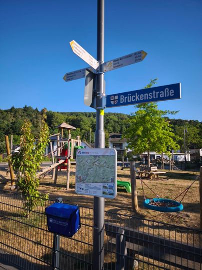 A traffic sign at a playground intersection shows the way to Brückenstraße. In the background, playground equipment and trees can be seen.