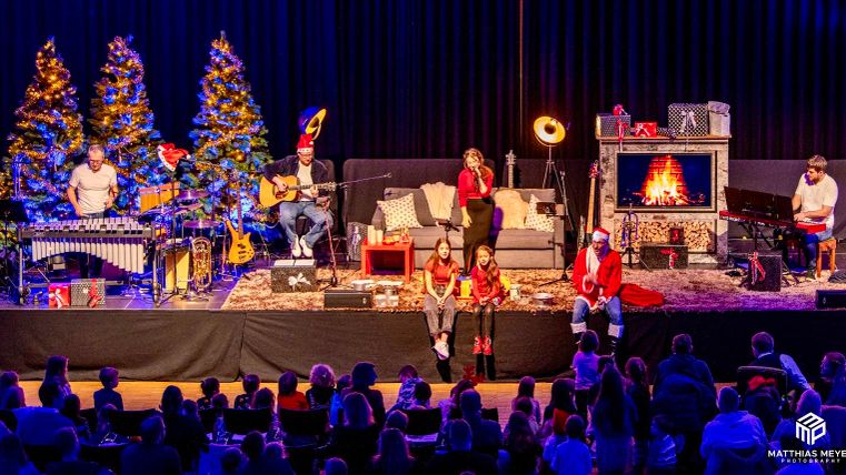 Une représentation festive de Noël avec des musiciens sur scène. Des sapins de Noël sont en arrière-plan et un coin feu crée une ambiance chaleureuse.
