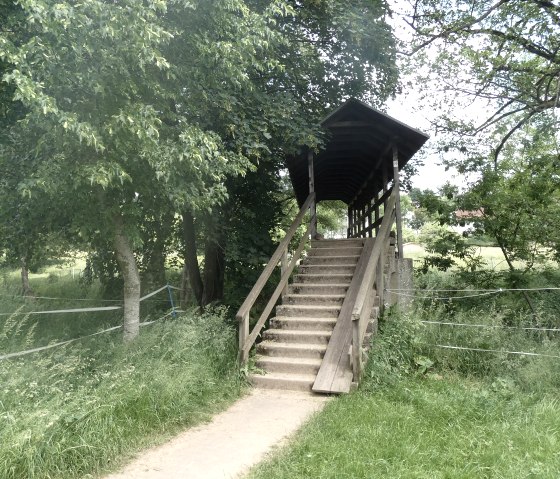 Überdachte Holzbrücke über die Nims, umgeben von grünen Bäumen und Wiesen., © TI Bitburger Land