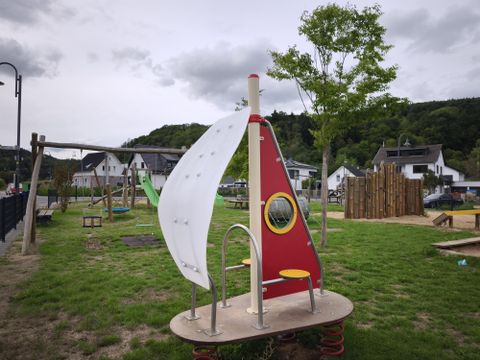 Een speeltuin met een speeltoestel in de vorm van een zeilboot en een rond raam. Op de achtergrond zijn bomen en woonhuizen te zien.
