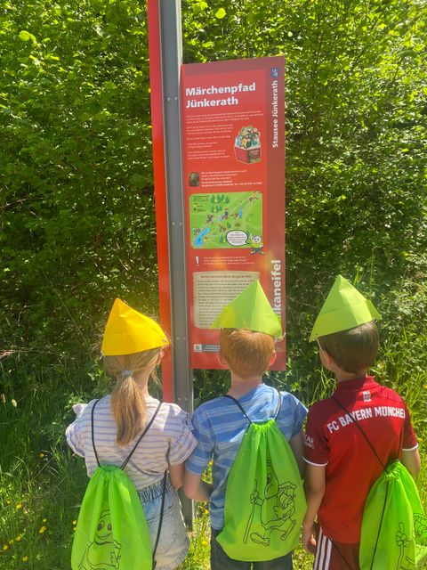 Drei Kinder mit bunten Hüten und grünen Rucksäcken stehen vor einem Schild des Märchenpfads Jünkerath.