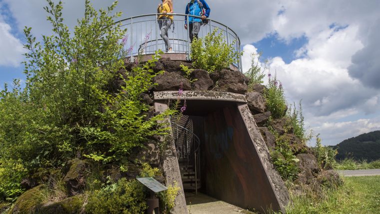 Eine Aussichtsplattform aus Stein mit zwei Personen darüber. Umgeben von grünen Pflanzen und einem hellen, bewölkten Himmel.