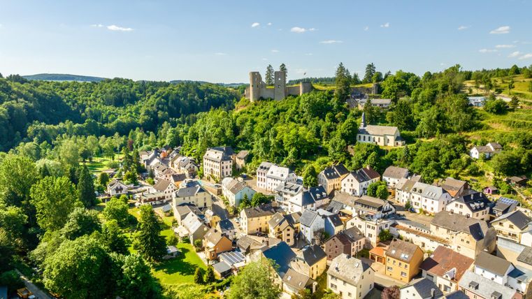 Eine malerische Landschaft mit einem kleinen Dorf und einer Burg auf einem Hügel. Umgeben von grünen Wäldern und blauen Himmel.