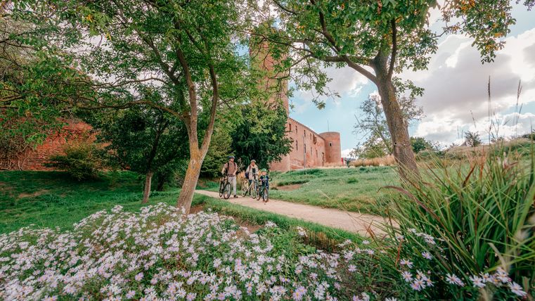 Three cyclists on a path in the park with a view of Zülpich Castle, surrounded by trees and flowers.