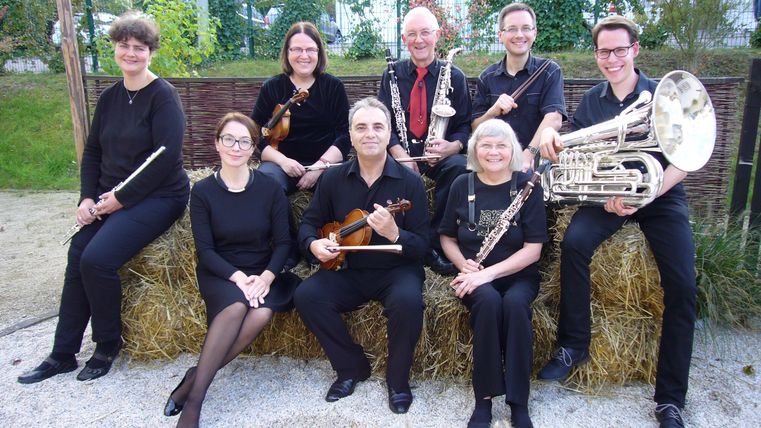 Eine Gruppe von Musikern sitzt auf einem Heuballen. Sie sind in schwarzen Outfits gekleidet und halten verschiedene Musikinstrumente.