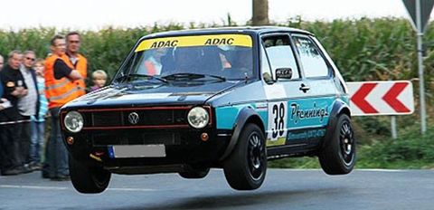 A sporty Volkswagen hovering in the air as it takes a turn. In the background, spectators and green countryside are visible.