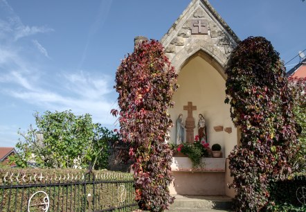 Een kleine kapel in Wolsfeld, omgeven door rode klimplanten. Binnen zijn religieuze beelden te zien. De lucht is helder en blauw., © Tourist-Information Bitburger Land