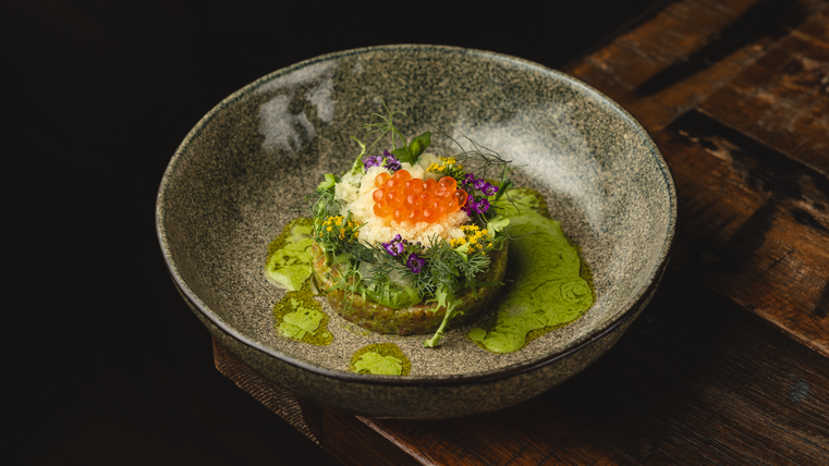 Une assiette artistiquement dressée avec des herbes fraîches, des fleurs comestibles colorées et du caviar de poisson orange. L'arrière-plan est sombre, et l'assiette se trouve sur une table en bois rustique.