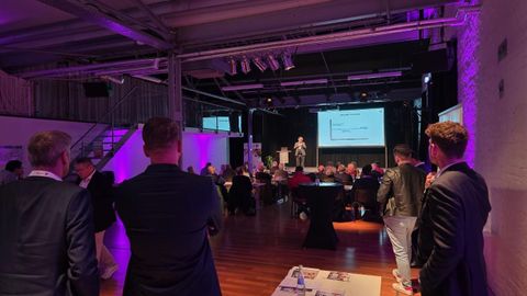 A conference with participants listening to a speaker on stage. In the background, presentation slides are visible on a large screen.