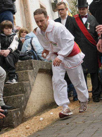 Ein Mann in einer weißen Tracht läuft eine Treppe hinunter und balanciert dabei Eier. Um ihn herum schauen Zuschauer gespannt zu.