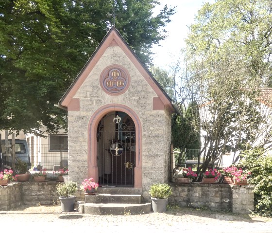 Petite chapelle à Idenheim, entourée de fleurs et d'arbres. Un panneau de signalisation indique 'Meilbrücker Straße'. La chapelle a un toit pointu et une porte grillagée., © TI Bitburger Land