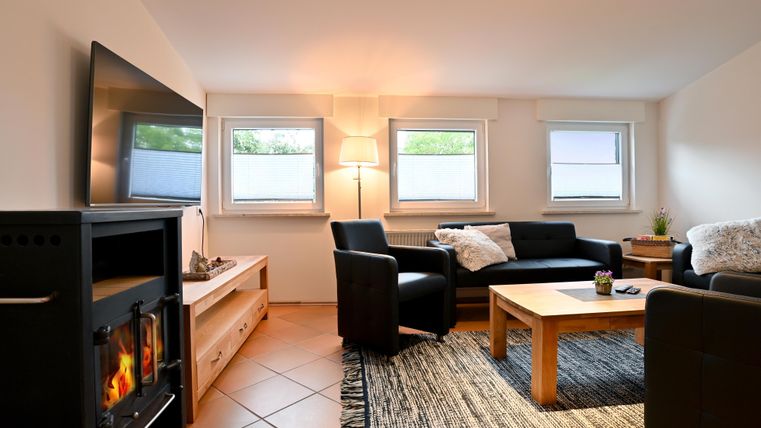 Ein gemütliches Wohnzimmer mit einem modernen Fernseher, bequemen Sofas und einem Tisch. Die großen Fenster sorgen für viel Tageslicht.