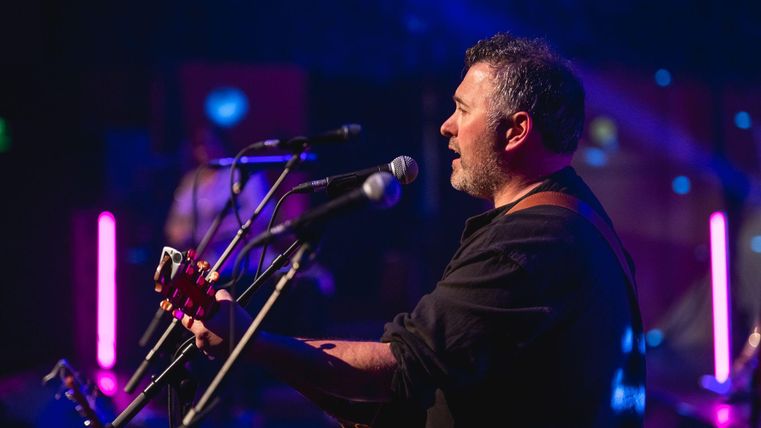 Ein Musiker steht auf der Bühne und spielt Gitarre. Im Hintergrund sind bunte Lichter und weitere Musiker zu sehen.