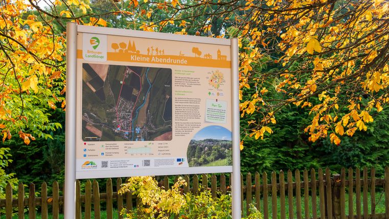 Informationsschild zur Wanderroute 'Kleine Abendrunde' im Bitburger Land, umgeben von herbstlichen Bäumen.