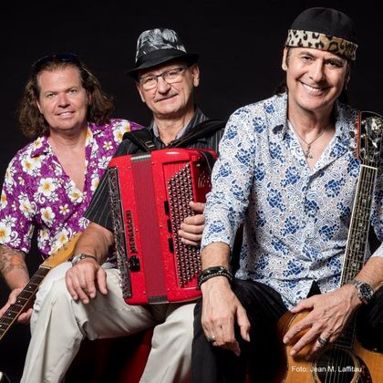 Three musicians are posing together in front of a black background. They are wearing colorful shirts and holding various musical instruments in their hands.