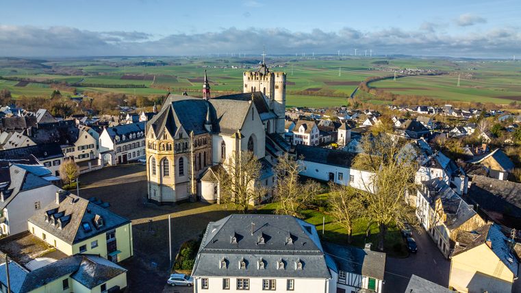Luchtfoto van Münstermaifeld met de stiftskerk en omliggende gebouwen, weidse velden en windturbines op de achtergrond.