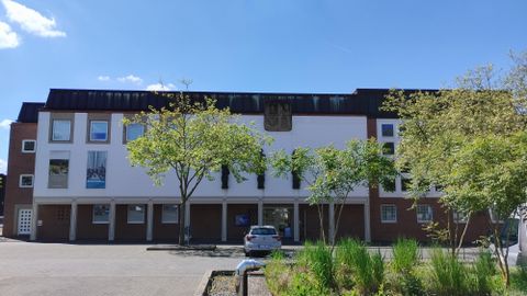 Ein modernes Gebäude mit einer hellen Fassade und großen Fenstern. Vor dem Gebäude stehen einige Bäume und es gibt einen Parkplatz.