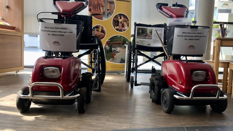 Deux fauteuils roulants motorisés rouges sont côte à côte dans un intérieur. À l'arrière-plan, des affiches d'information sont visibles.